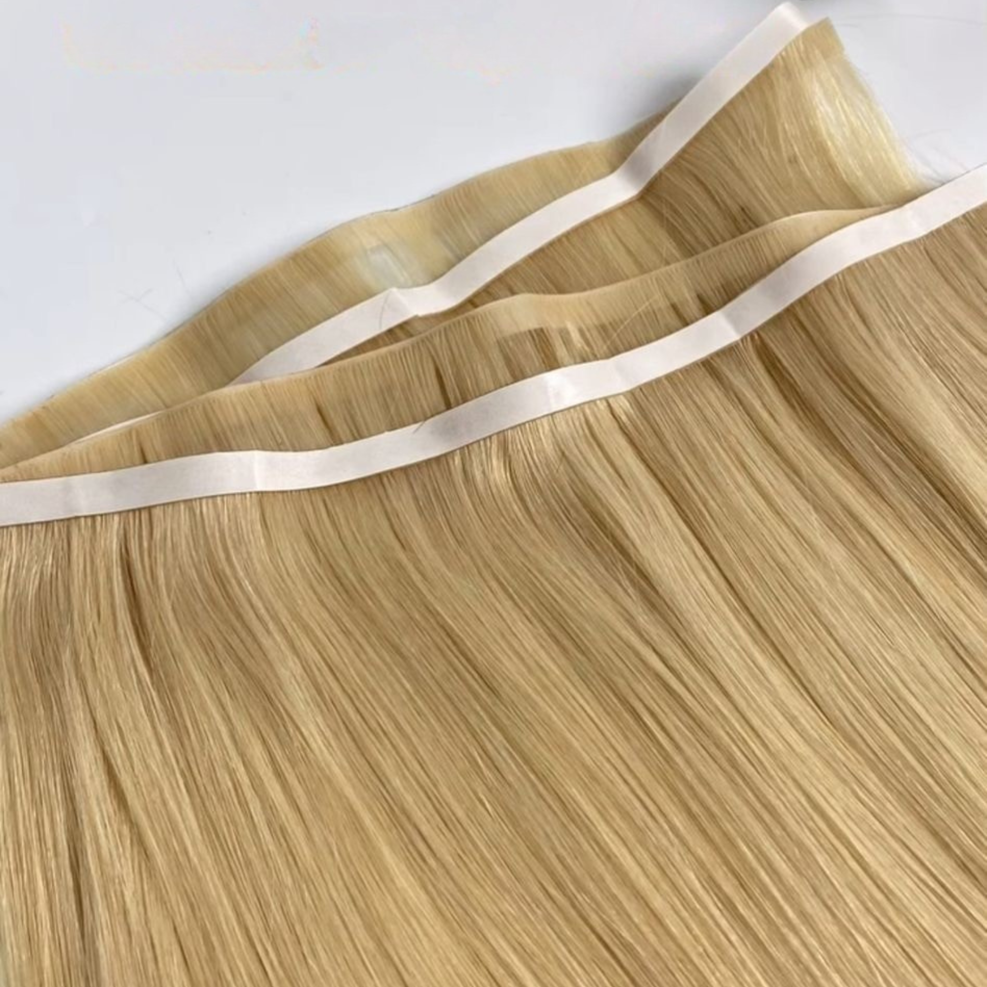Close-up of platinum blonde invisible tape weft hair extensions with ultra-flat adhesive tape sections visible along the top. The silky Remy human hair has a smooth, glossy finish and showcases the seamless tape weft construction against a clean white background