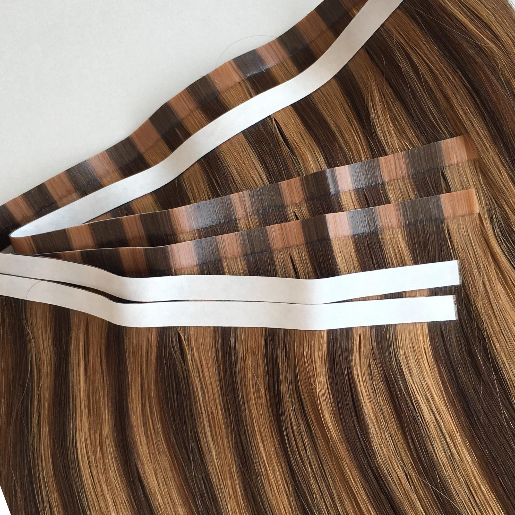 Close-up of dimensional brunette and blonde tape weft hair extensions with wide adhesive tape sections visible along the top. The blended Remy human hair features soft caramel and blonde tones with a seamless tape weft design and glossy finish against a clean white background.